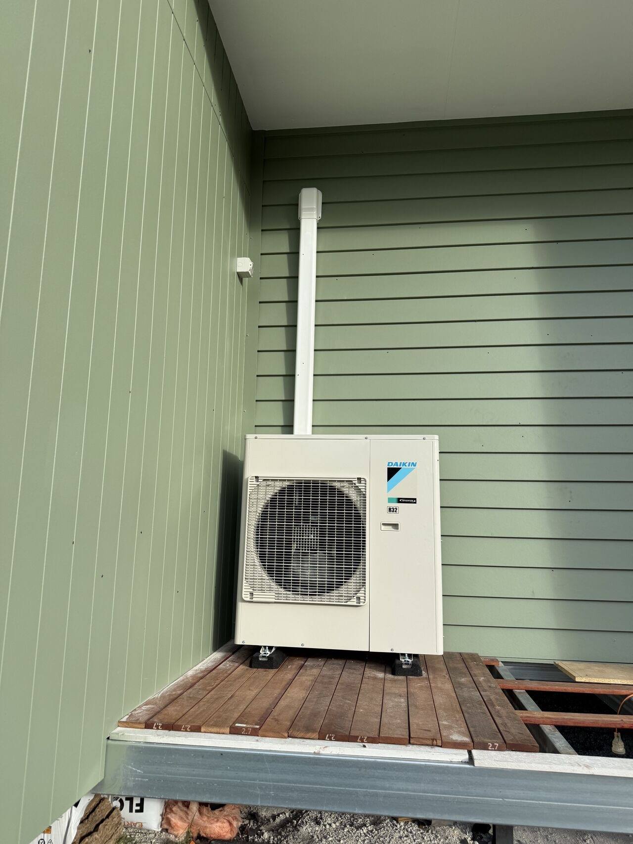 Outdoor condenser on a raised deck platform beside green weatherboard cladding