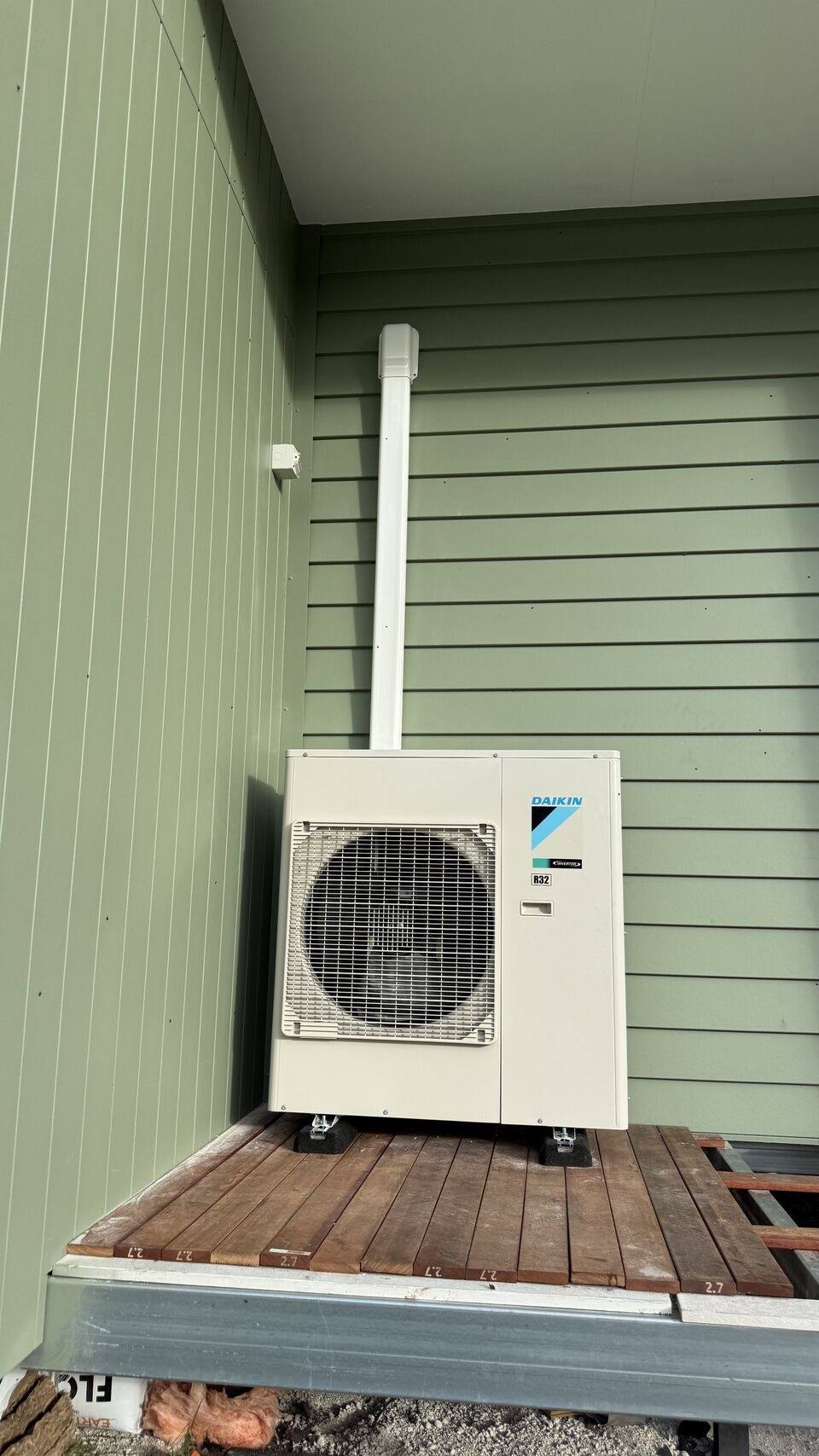 Outdoor condenser on a raised deck platform beside green weatherboard cladding